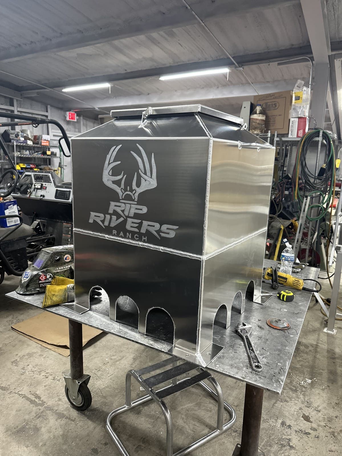 Aluminum quail feeder on legs with a laser-cut ranch logo and arched feed openings around the base.